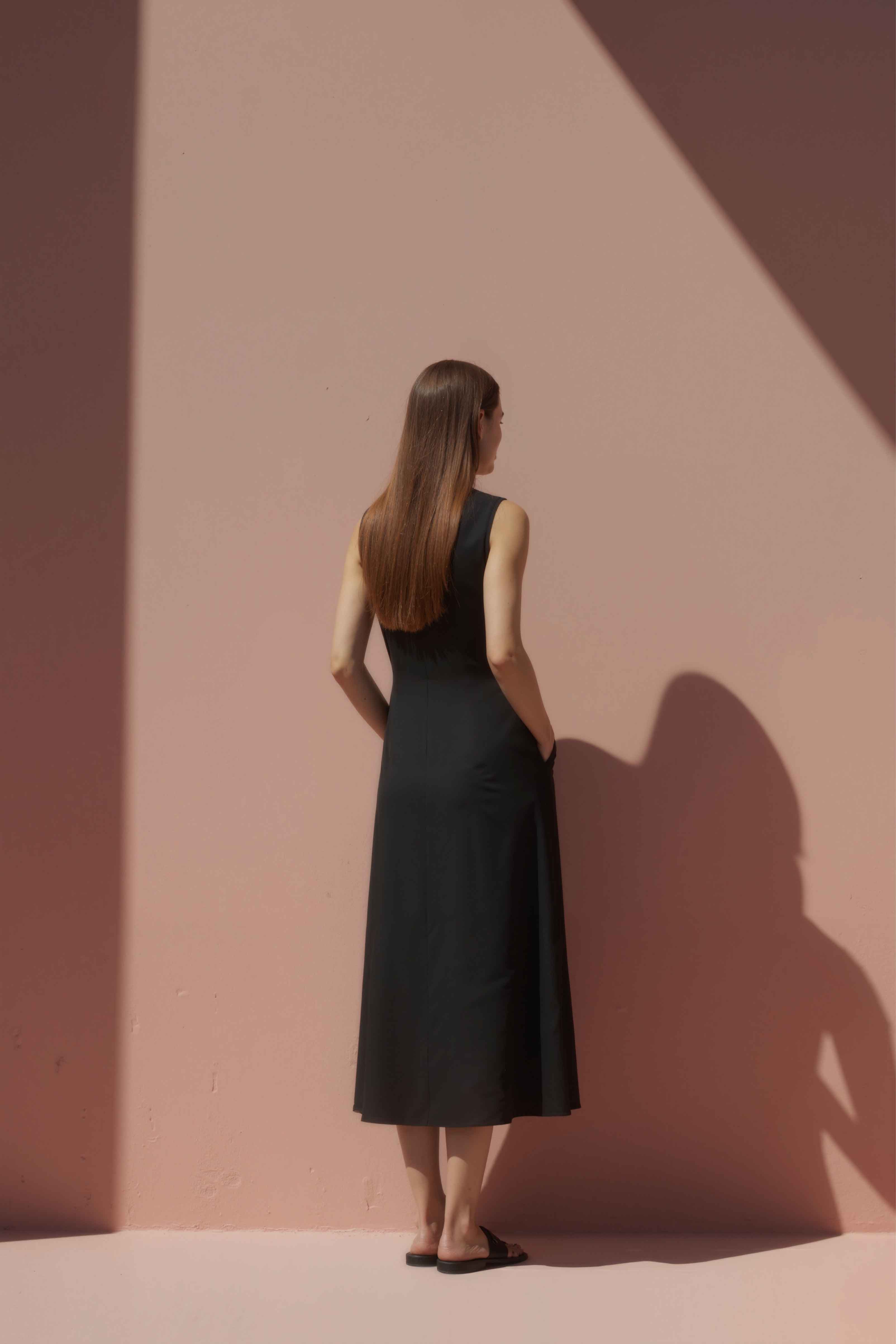 Woman in a black cotton shift dress standing against a pink wall.