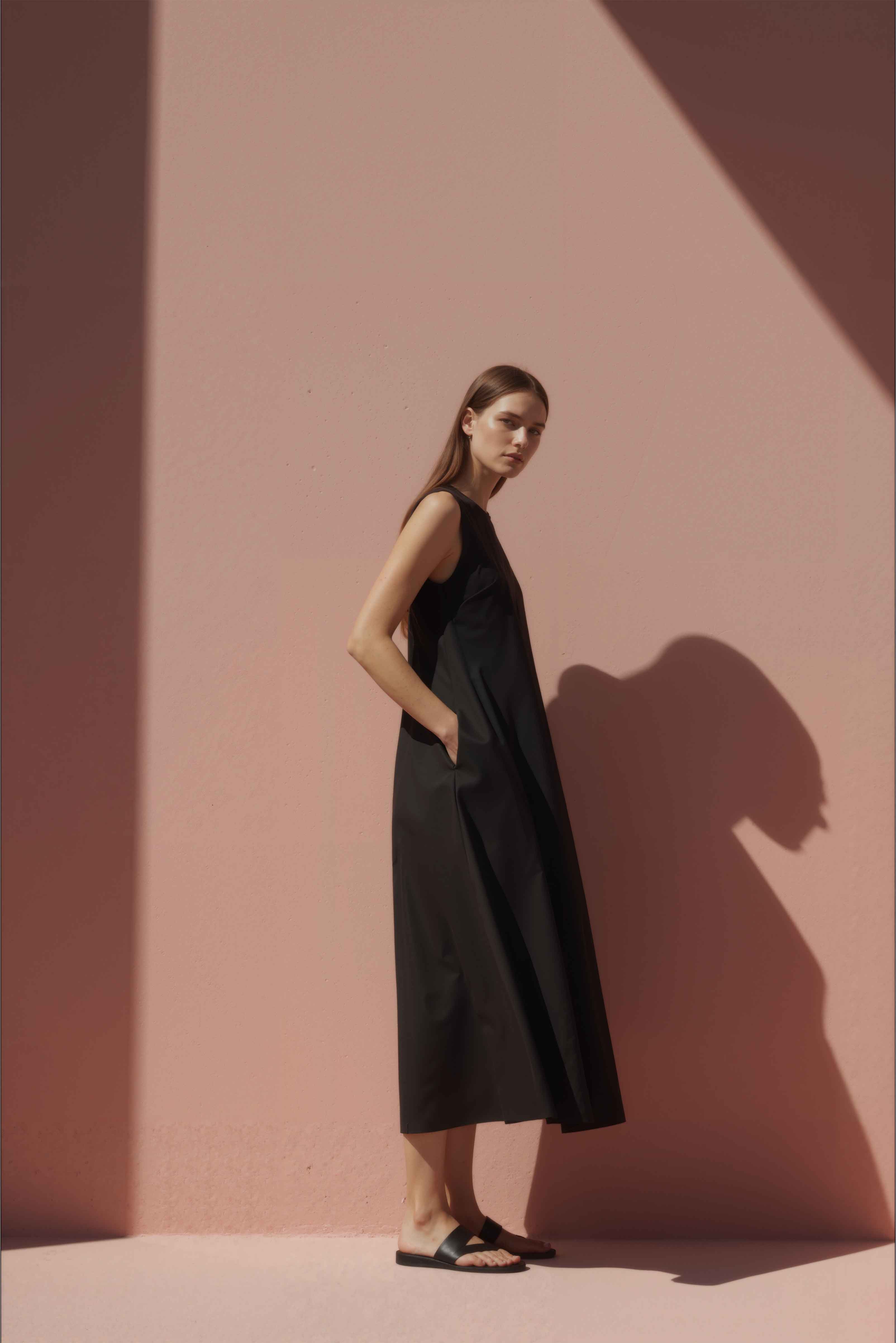 Woman in a black cotton shift dress standing against a pink wall.