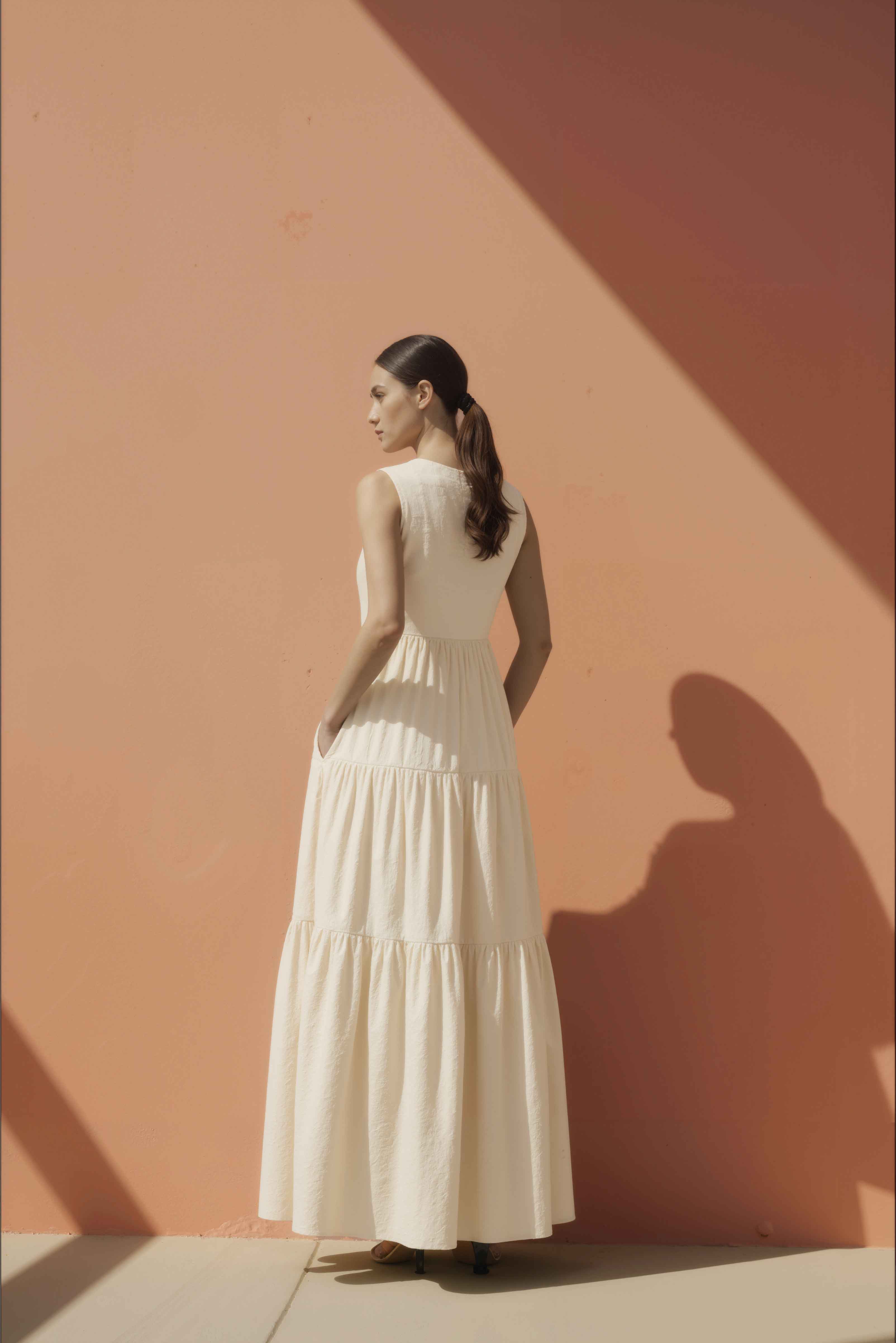 Woman in a white cotton maxi dress standing against a pink wall.