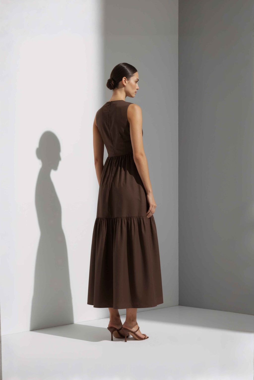Anasua The Forme Edit Poplin Cotton Midi Dress in Umber Brown, a structured sleeveless cotton midi dress with a tiered skirt, photographed under natural sunlight.