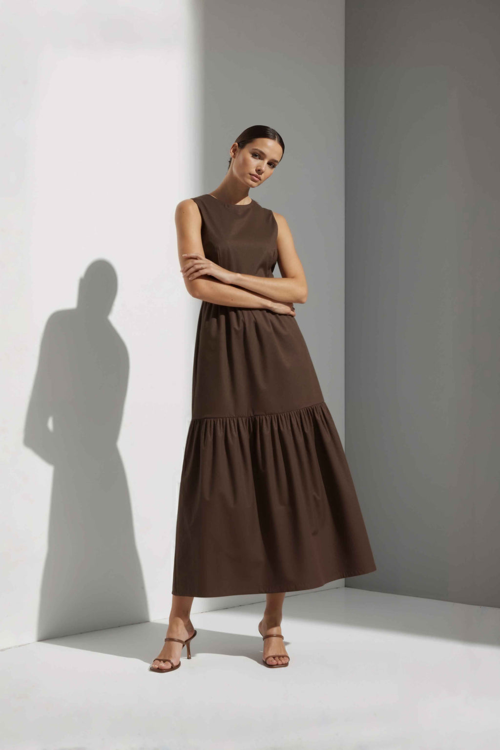 Anasua The Forme Edit Poplin Cotton Midi Dress in Umber Brown, a structured sleeveless cotton midi dress with a tiered skirt, photographed under natural sunlight.