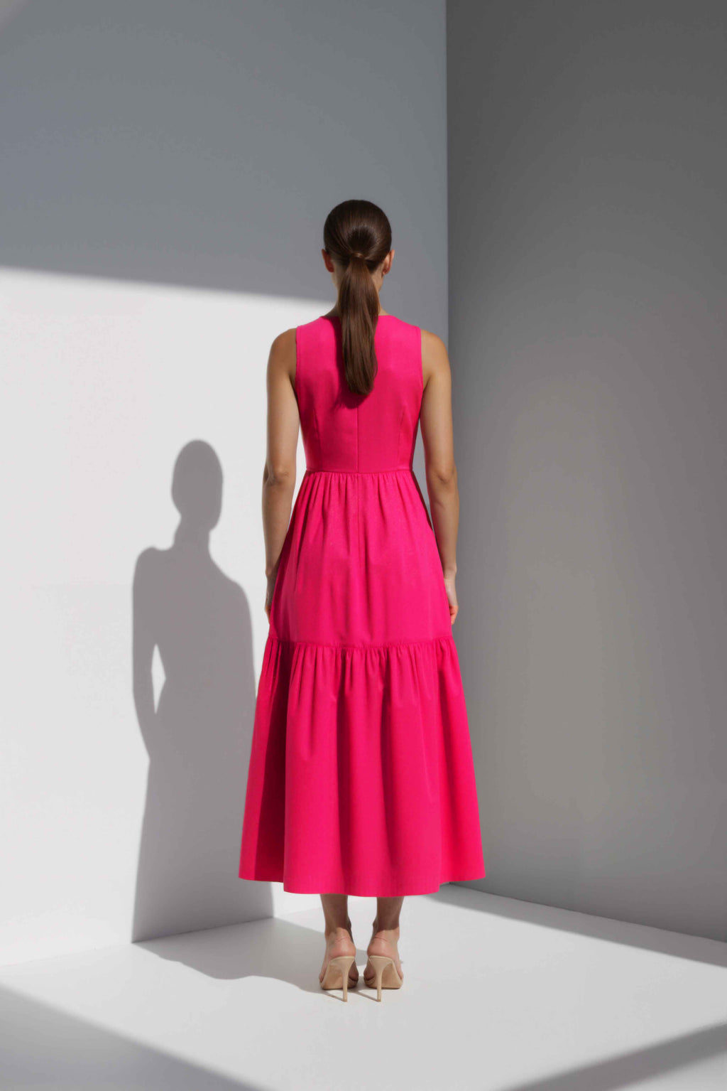 Model wearing The Forme Edit Poplin Cotton Midi Dress in Pink, photographed under natural sunlight against a minimal architectural wall, highlighting the structured bodice and softly tiered skirt.