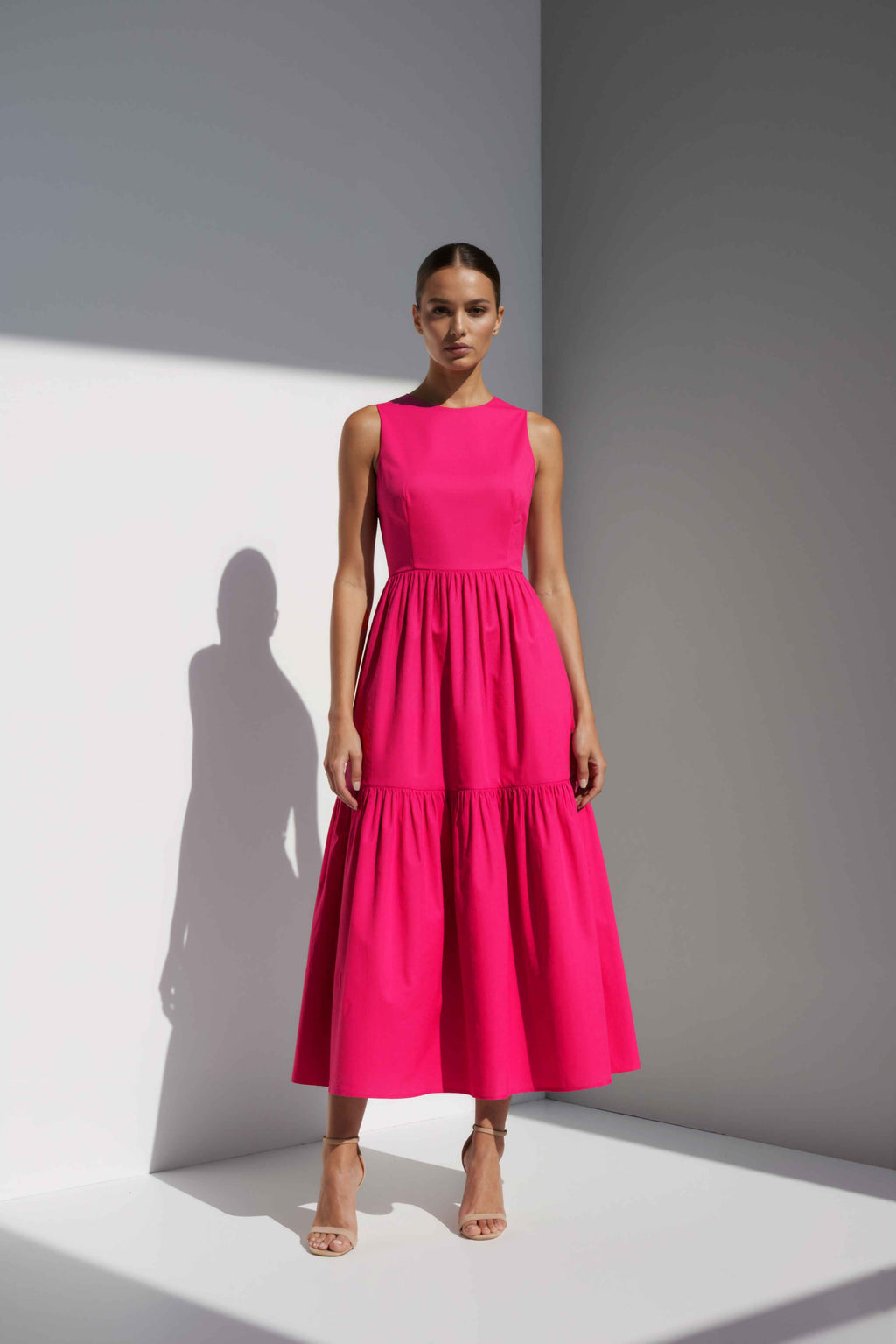 Model wearing The Forme Edit Poplin Cotton Midi Dress in Pink, photographed under natural sunlight against a minimal architectural wall, highlighting the structured bodice and softly tiered skirt.