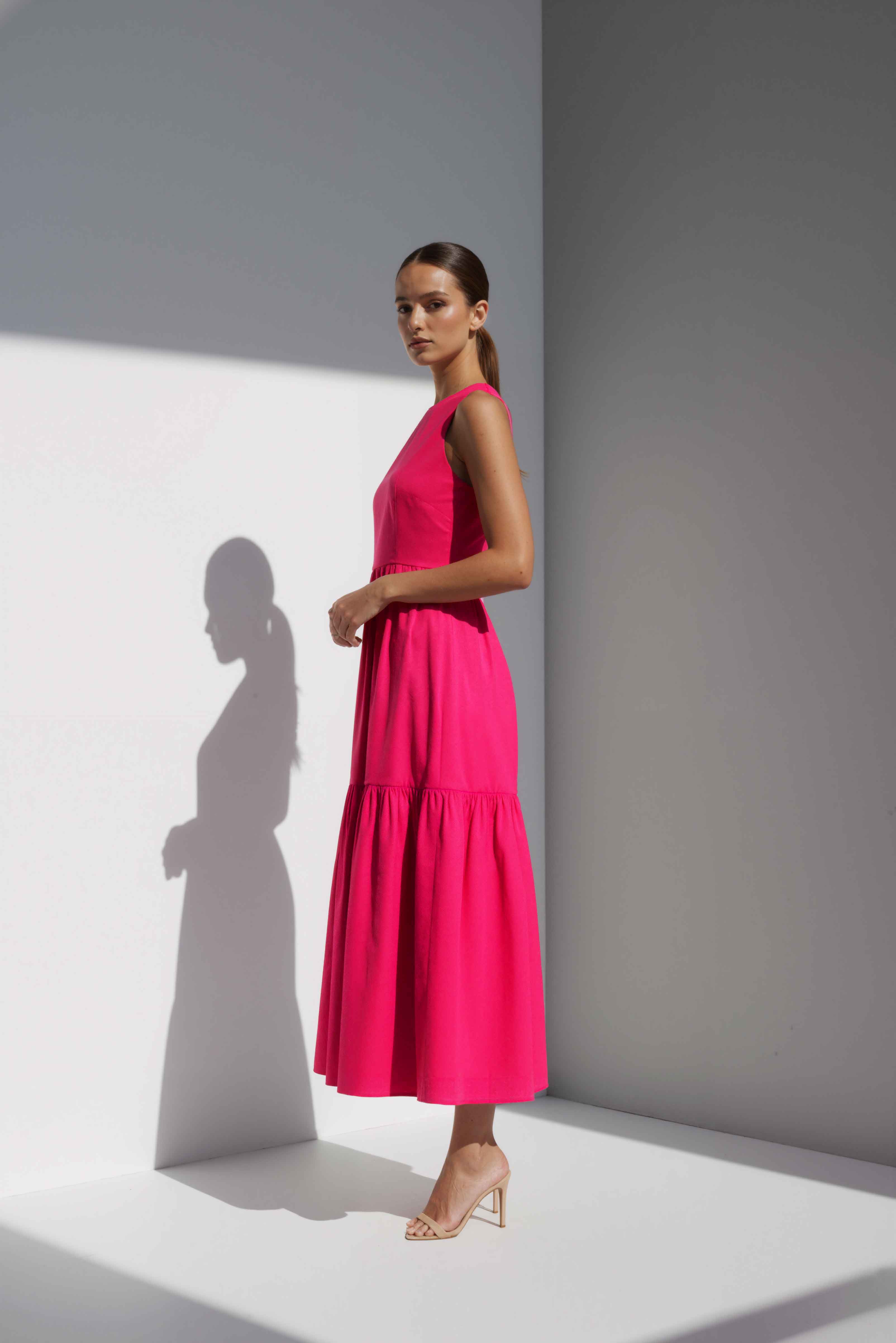 Model wearing The Forme Edit Poplin Cotton Midi Dress in Pink, photographed under natural sunlight against a minimal architectural wall, highlighting the structured bodice and softly tiered skirt.