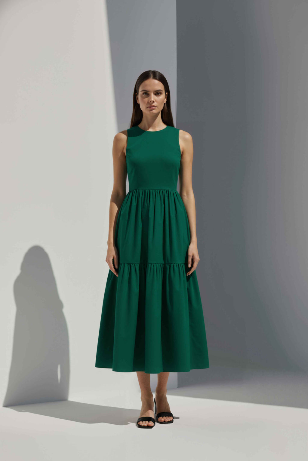 Anasua The Forme Edit Poplin Cotton Midi Dress in Verdant Green, a structured sleeveless cotton midi dress with a tiered skirt, photographed under natural sunlight.