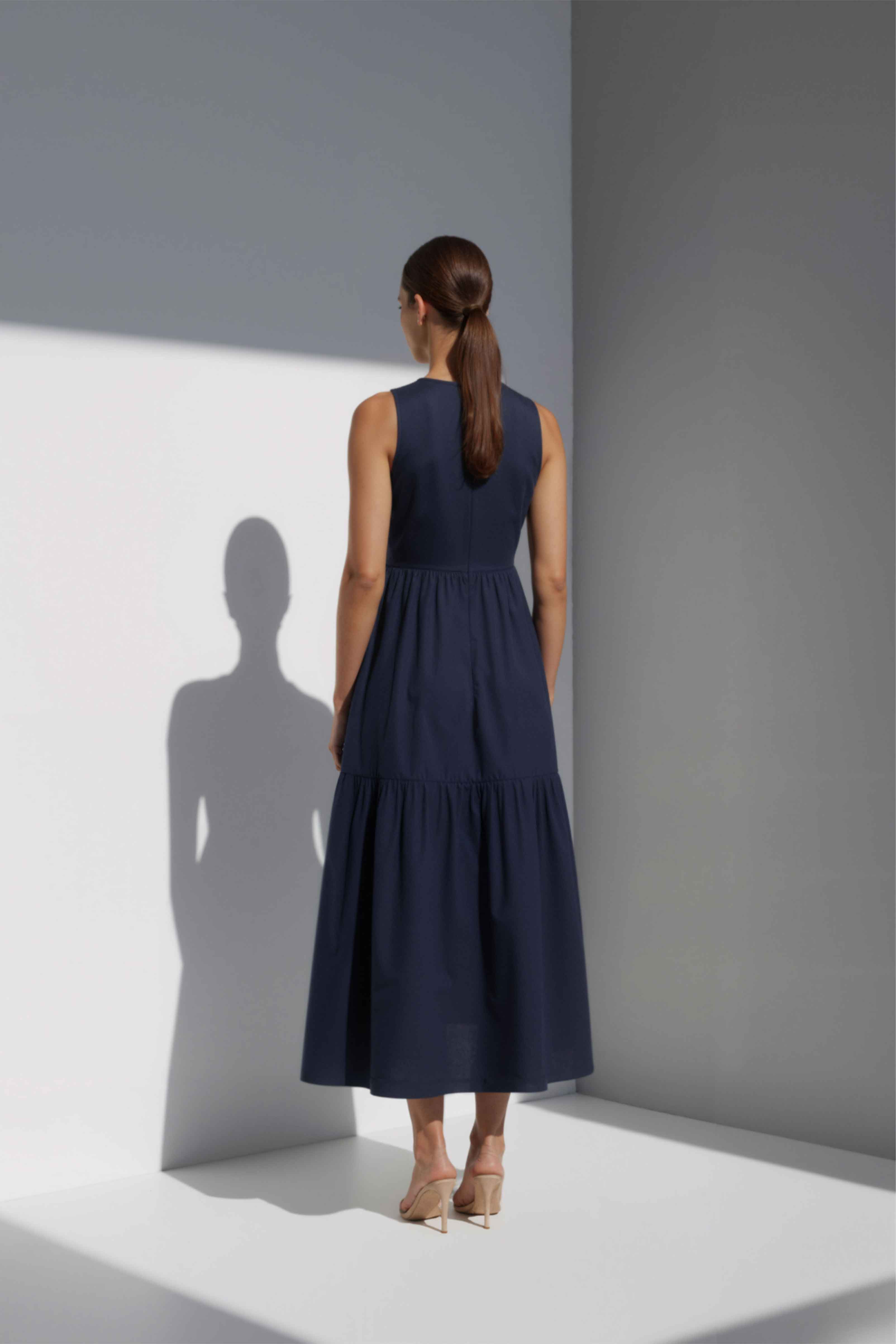 Model wearing The Forme Edit Poplin Cotton Midi Dress in Navy Blue, photographed under natural sunlight against a minimal architectural wall, highlighting the structured bodice and softly tiered skirt.
