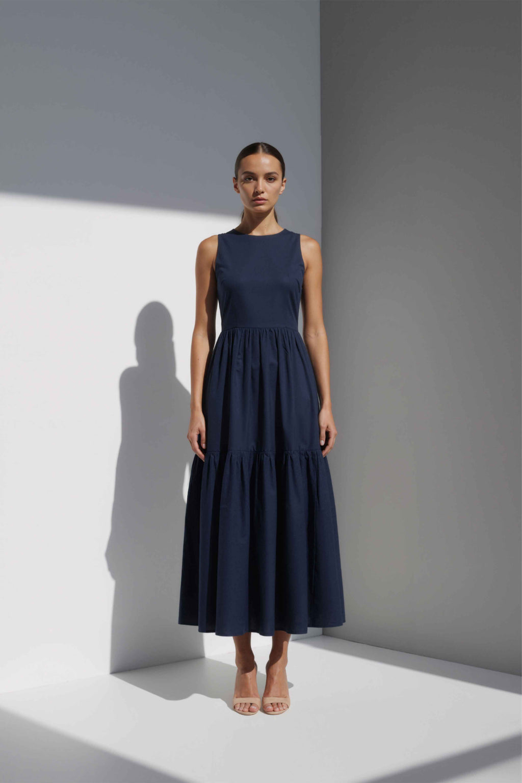 Model wearing The Forme Edit Poplin Cotton Midi Dress in Navy Blue, photographed under natural sunlight against a minimal architectural wall, highlighting the structured bodice and softly tiered skirt.