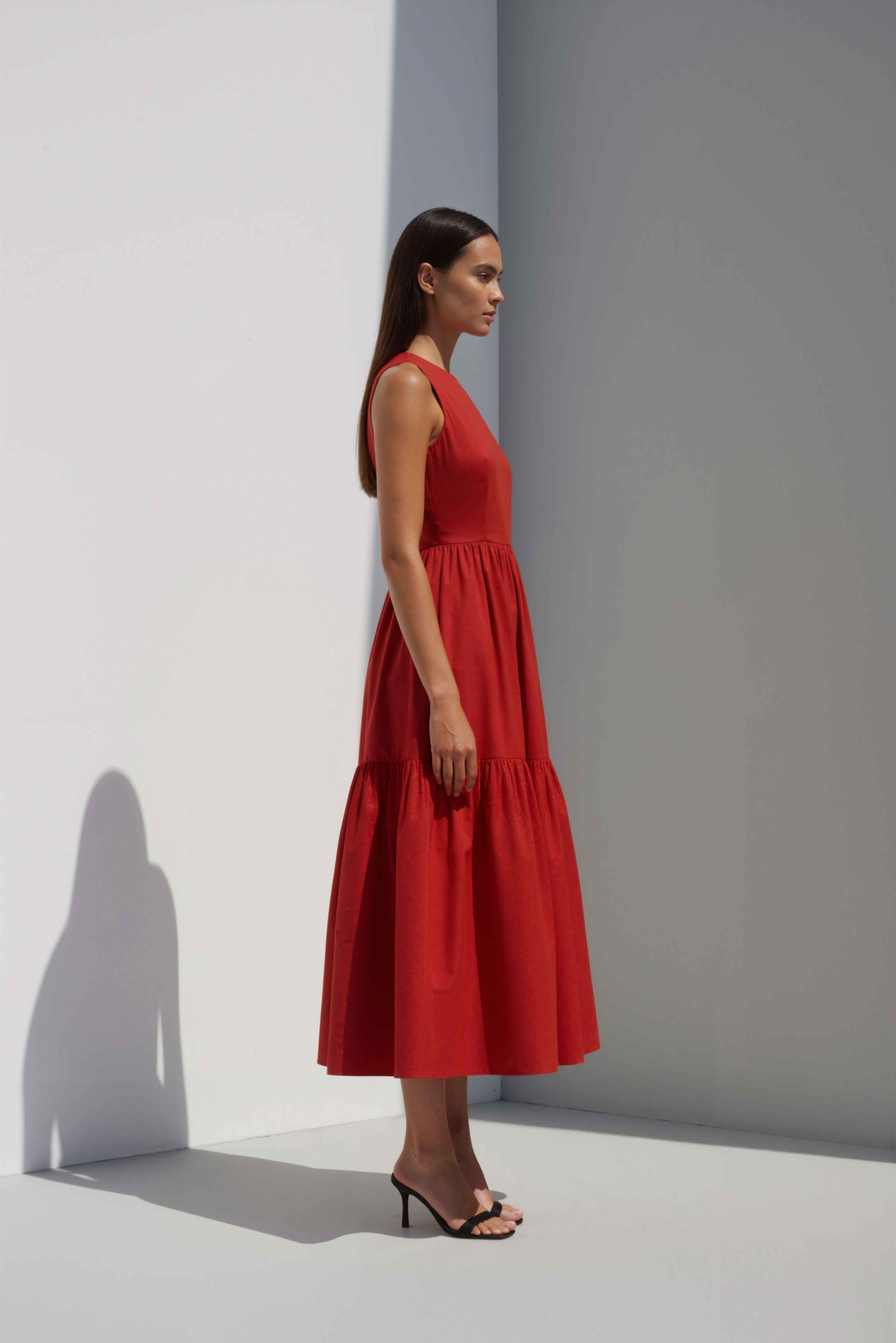 Anasua The Forme Edit Poplin Cotton Midi Dress in Sienna Red, a structured sleeveless cotton midi dress with a tiered skirt, photographed under natural sunlight.