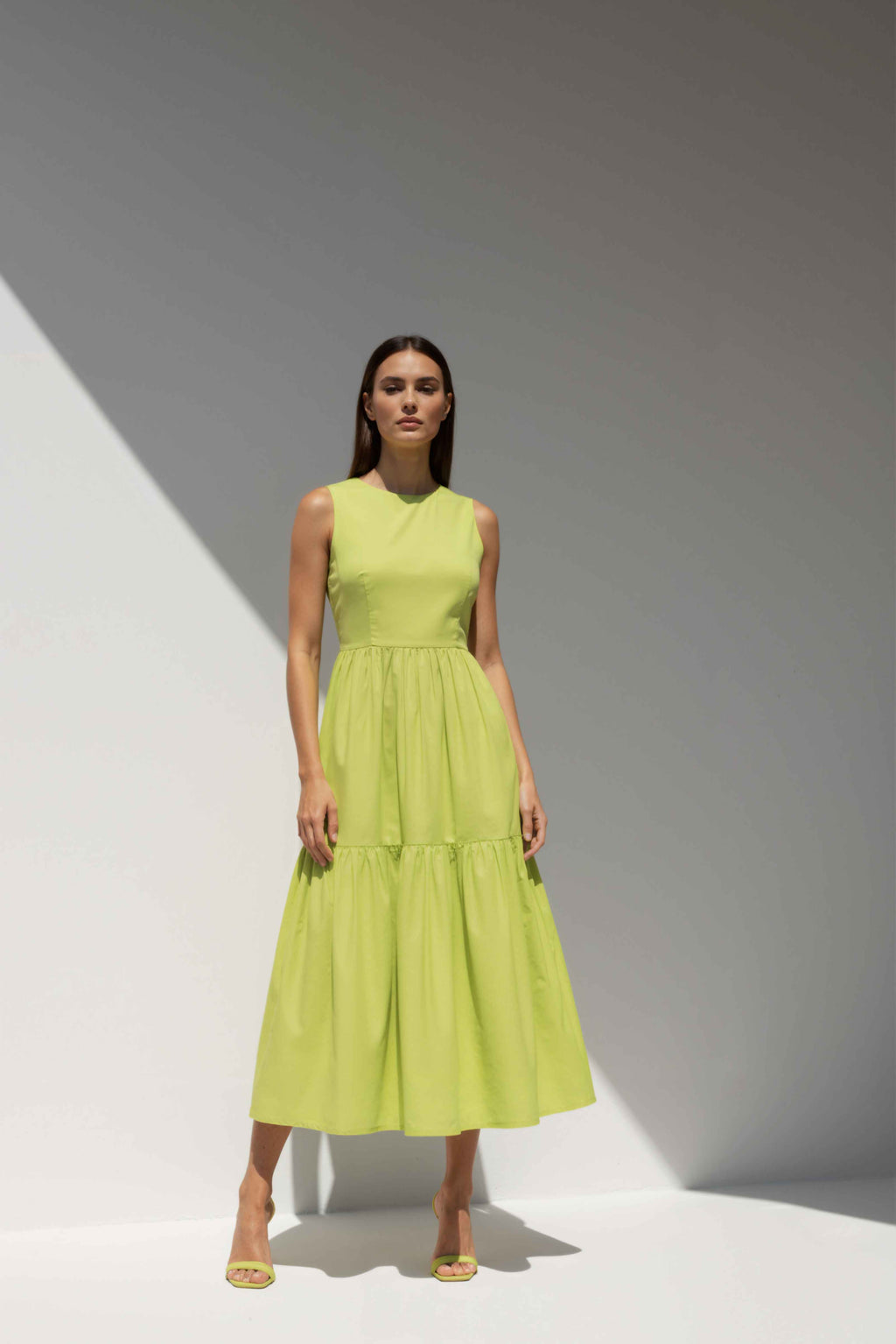 Model wearing The Forme Edit Poplin Cotton Midi Dress in Yellow, photographed under natural sunlight against a minimal architectural wall, highlighting the structured bodice and softly tiered skirt.